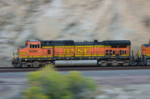 AN ORANGE, YELLOW AND BLACK NBSF-LABELED LOCOMOTIVE IS SPEEDING PASSED A CANYON WITH A SINGLE TREE IN THE FOREGROUND OF THE IMAGE.