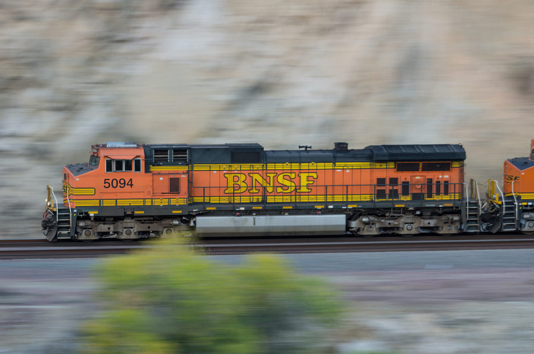AN ORANGE, YELLOW AND BLACK NBSF-LABELED LOCOMOTIVE IS SPEEDING PASSED A CANYON WITH A SINGLE TREE IN THE FOREGROUND OF THE IMAGE.