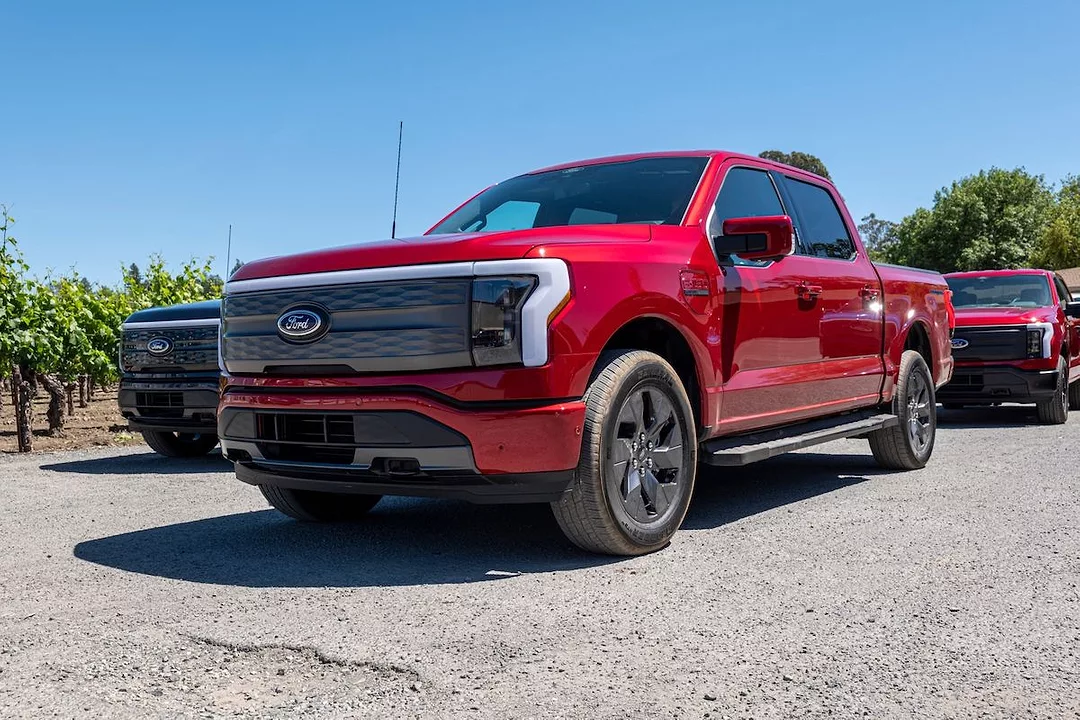 A RED FORD LIGHTNING F-150 IS PARKED NEAR SOME TREES AND OTHER TRUCKS ON A CLEAR, SUNNY DAY.