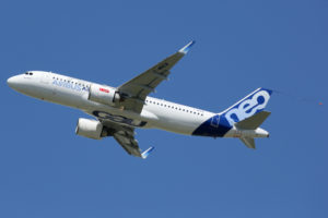 A WHITE AIRBUS-LABELED AIRPLANE IS FLYING THROUGH THE SKY. THE BLUE RUDDER HAS THE WORDS "NEO" WRITTEN ON IT IN WHITE LETTERING. NEO IS WRITTEN UNDERNEATH THE BOTTOM OF THE PLANE IN BLUE.