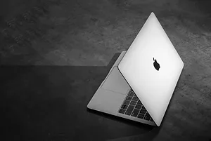 A PARTIALLY-OPENED WHITE APPLE LAPTOP SITS ON A DARK FLOOR.