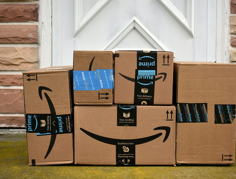 SEVERAL DIFFERENT-SIZED AMAZON BOXES SIT IN FRONT OF A WHITE DOOR SURROUNDED BY BRICKS.