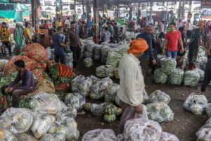 MANY PEOPLE CAN BE SEEN HOLDING OR WALKING NEAR PLASTIC BAGS FULL OF VEGETABLES IN A COVERED MARKET.