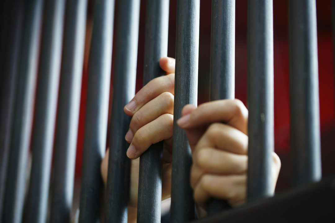 A CLOSE-UP OF A PAIR OF HANDS HOLDS ONTO PRISON CELL BARS.