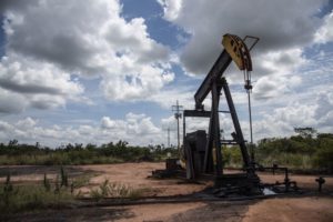 A PUMP JACK IS NEAR AN OIL SPILL IN A SMALL, DUSTY FIELD SURROUNDED BY TREES.