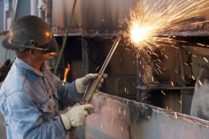 A MAN IN BLUE OVERALLS AND PROTECTIVE MASK WELDS PIECES OF A SHIP, SPARKS FLYING
