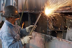 A MAN IN BLUE OVERALLS AND PROTECTIVE MASK WELDS PIECES OF A SHIP, SPARKS FLYING
