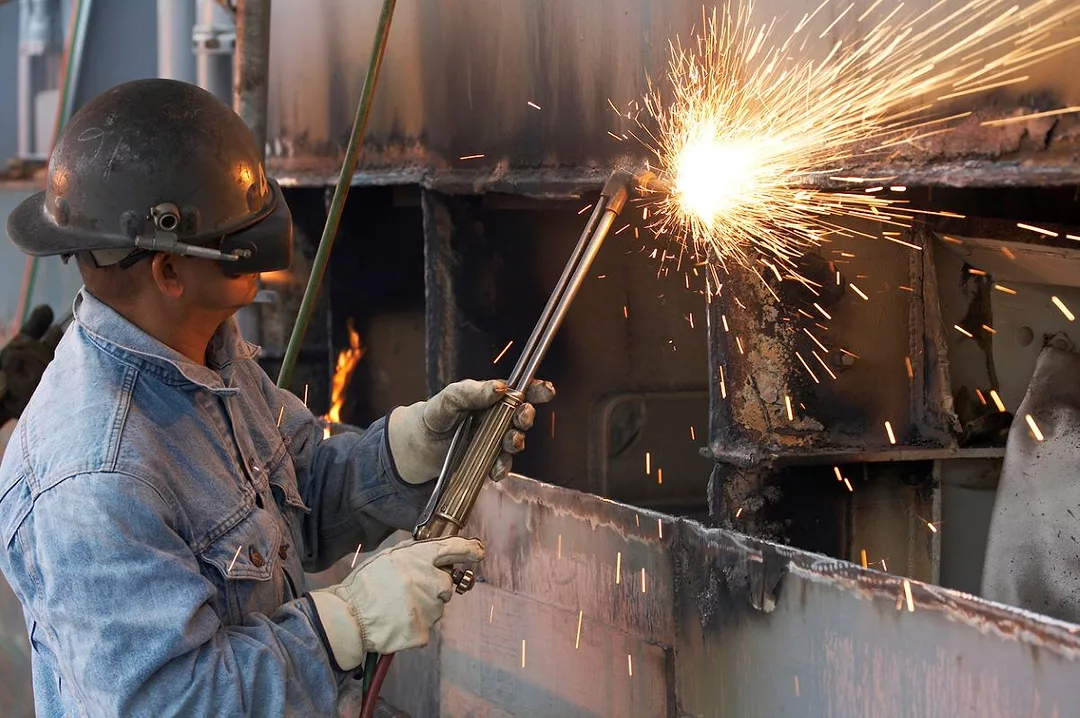 A MAN IN BLUE OVERALLS AND PROTECTIVE MASK WELDS PIECES OF A SHIP, SPARKS FLYING