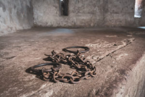 RUSTED HANDCUFFS SIT ON THE FLOOR OF AN OLD, DINGY HOLDING CELL.