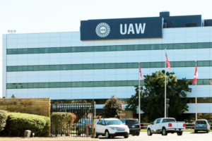 CARS ARE DRIVING IN AND OUT OF THE GATED ENTRANCE OF THE UNITED AUTO WORKERS' UNION HEADQUARTERS. A UAW SIGN HANGS ON THE FRONT OF THE BUILDING.