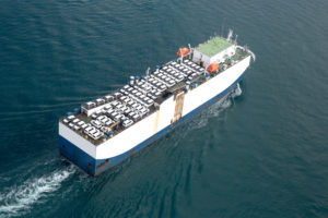 A TRANSPORT VESSEL CAN BE SEEN SAILING ON THE OCEAN WHILE CARRYING SEVERAL VEHICLES.