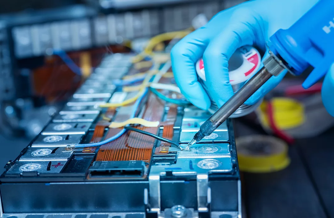 A GLOVED HAND USES A SOLDERING IRON ON A CAR BATTERY.