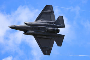 THE UNDERSIDE OF AN F-35 LIGHTNING FIGHTER JET CAN BE SEEN IN FRONT OF A PARTLY CLOUDY SKY.