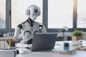A HUMANOID ROBOT WEARING A PAIR OF WHITE HEADPHONES SITS AT A DESK IN FRONT OF AN OPEN LAPTOP.