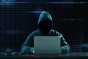 A HOODED FIGURE TYPES ON A COMPUTER, THEIR FACE OBSCURED IN DARKNESS