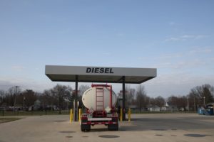 A TANKER TRUCK IS PARKED AT A DIESEL FUEL PUMP. THE WORD "DIESEL" IS WRITTEN ON THE PUMPING STATION.
