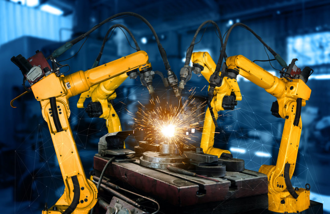 FOUR YELLOW ROBOTIC ARMS APPEAR TO BE WELDING A PIECE OF MACHINERY. SPARKS CAN BE SEEN COMING OFF THE MACHINE.