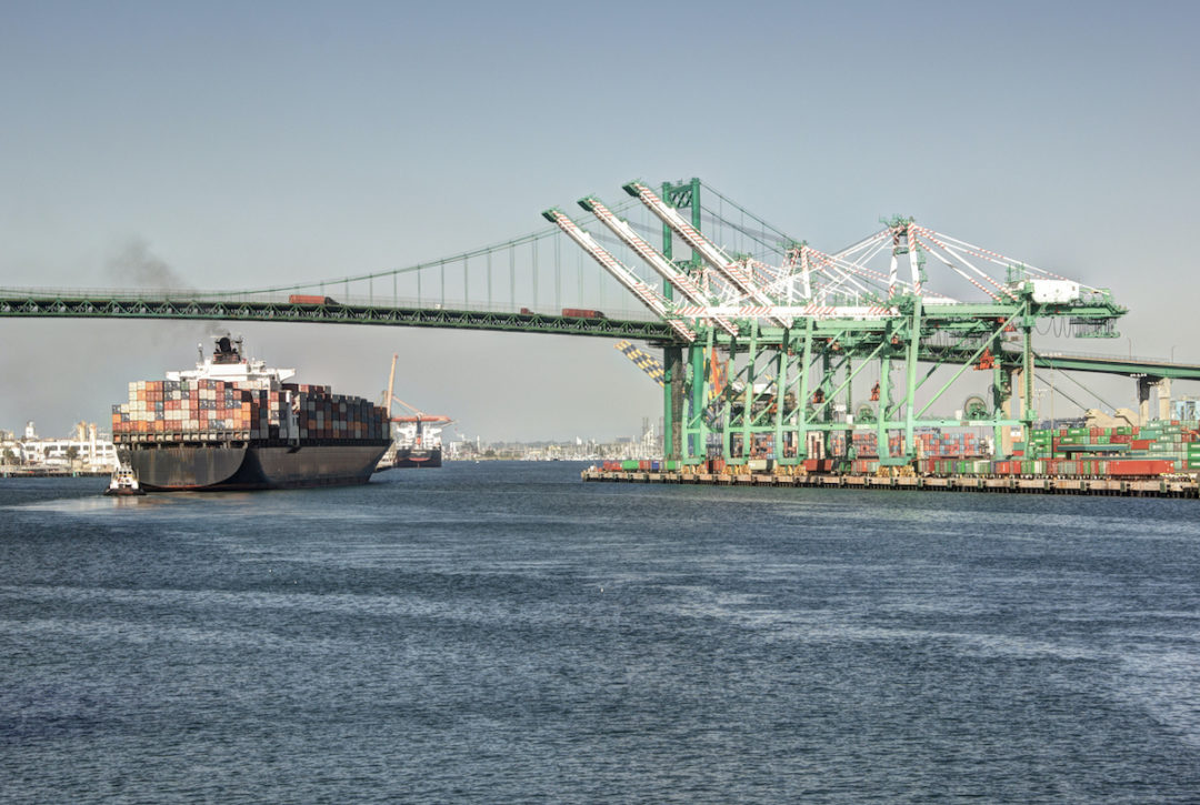 A LARGE CONTAINER SHIP SAILS UNDERNEATH A BRIDGE NEAR SOME CRANES AT A BUSY PORT.