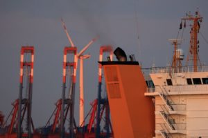THE ORANGE SMOKE STACK OF A LARGE SHIP STICKS OUT ABOVE A ROW OF PORT CRANES