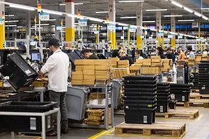 WORKERS STAND AT PICKING STATIONS IN A BUSY WAREHOUSE FULL OF PLASTIC BINS AND PACKAGES