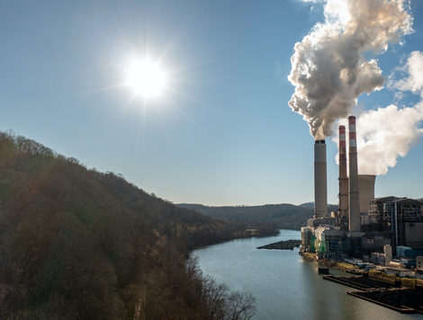 SMOKE BILLOWS OUT OF SMOKESTACKS AT A POWER PLANT LOCATED BY A BODY OF WATER. THE SUN SHINES DOWN ON THE PLANT.
