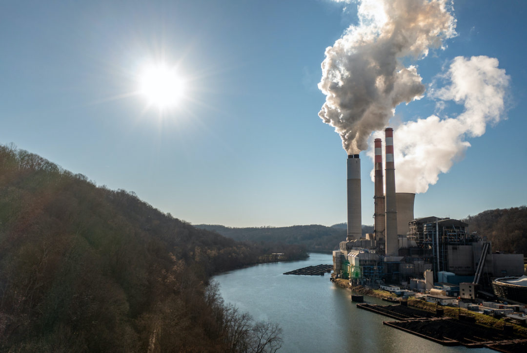 SMOKE BILLOWS OUT OF SMOKESTACKS AT A POWER PLANT LOCATED BY A BODY OF WATER. THE SUN SHINES DOWN ON THE PLANT.