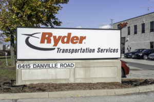 A RYDER SYSTEM INC SING SITS IN FRONT OF A FACILITY THAT HAS A RYDER LOGO ON IT.