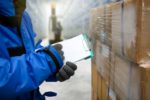 A WORKER IN COLD WEATHER GEAR WRITES ON A CLIPBOARD IN A WAREHOUSE