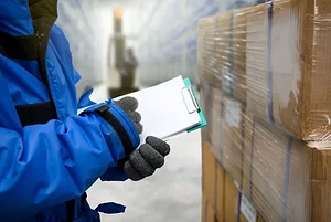 A WORKER IN COLD WEATHER GEAR WRITES ON A CLIPBOARD IN A WAREHOUSE