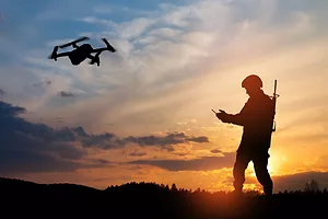 A SILHOUETTE OF A SOLDIER USING A REMOTE TO CONTROL A DRONE AT SUNSET.