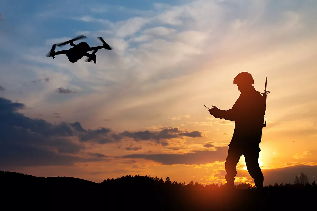 A SILHOUETTE OF A SOLDIER USING A REMOTE TO CONTROL A DRONE AT SUNSET.