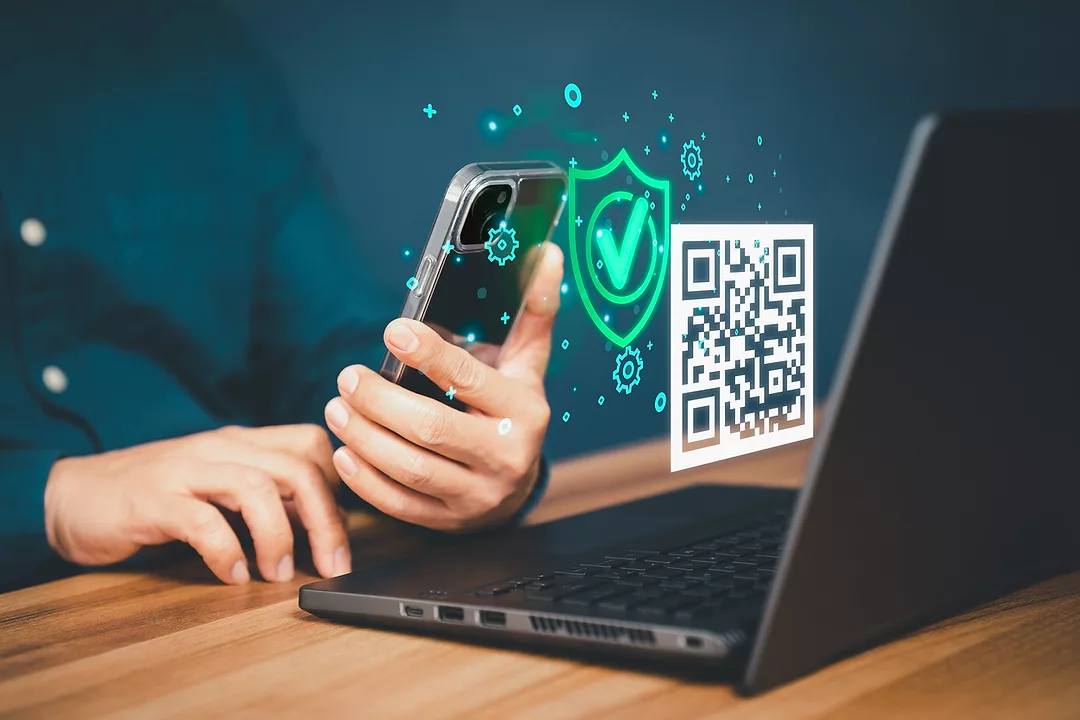 A HAND HOLDS A CELL PHONE IN FRONT OF A LAPTOP. A FLOATING QR CODE CAN BE SEEN BETWEEN THE PHONE AND LAPTOP.