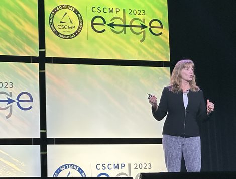 A WOMAN STANDS TALKING ON STAGE IN FRONT OF A CSCMP EDGE LOGO