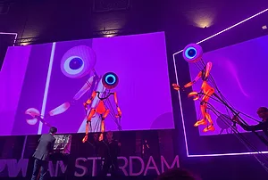 TALL, ILLUMINATED PUPPETS LIKE ROBOTS WITH A SINGLE GIANT EYE FOR A HEAD LOOM OVER A CONFERENCE STAGE