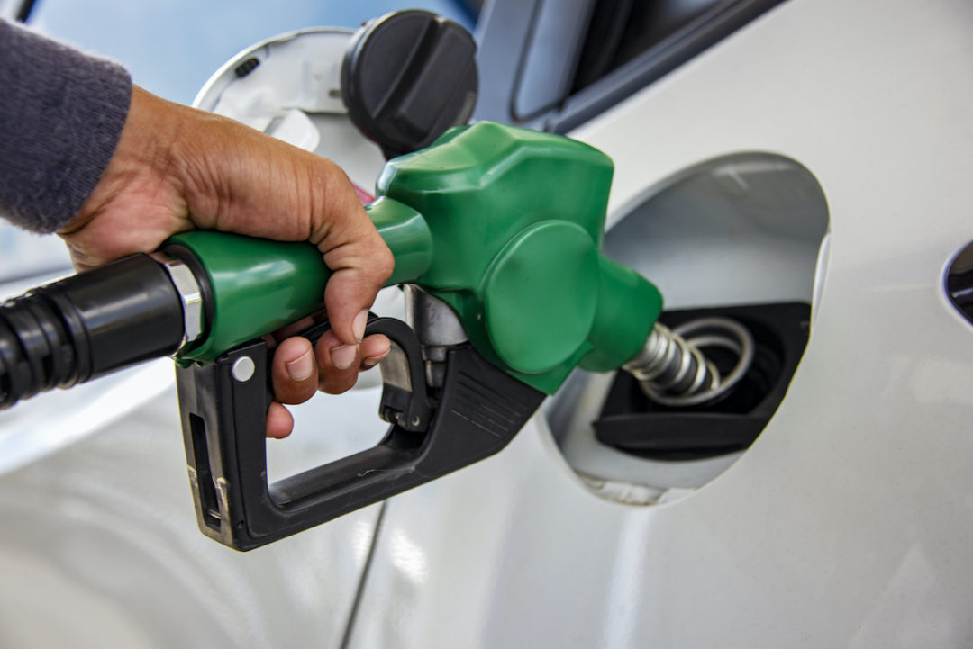 A PERSON'S HAND PLACES A GREEN GAS PUMP NOZZLE INTO THE GAS TANK OF A VEHICLE.