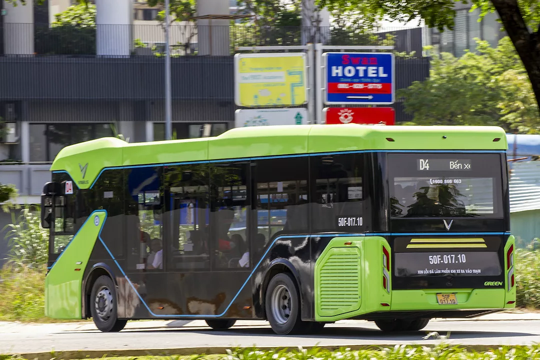 A GREEN AND BLACK ELECTRIC BUS DRIVES IN FRONT OF A SIGN. THE VINFAST LOGO CAN BE SEEN ON THE BACK OF THE BUS.