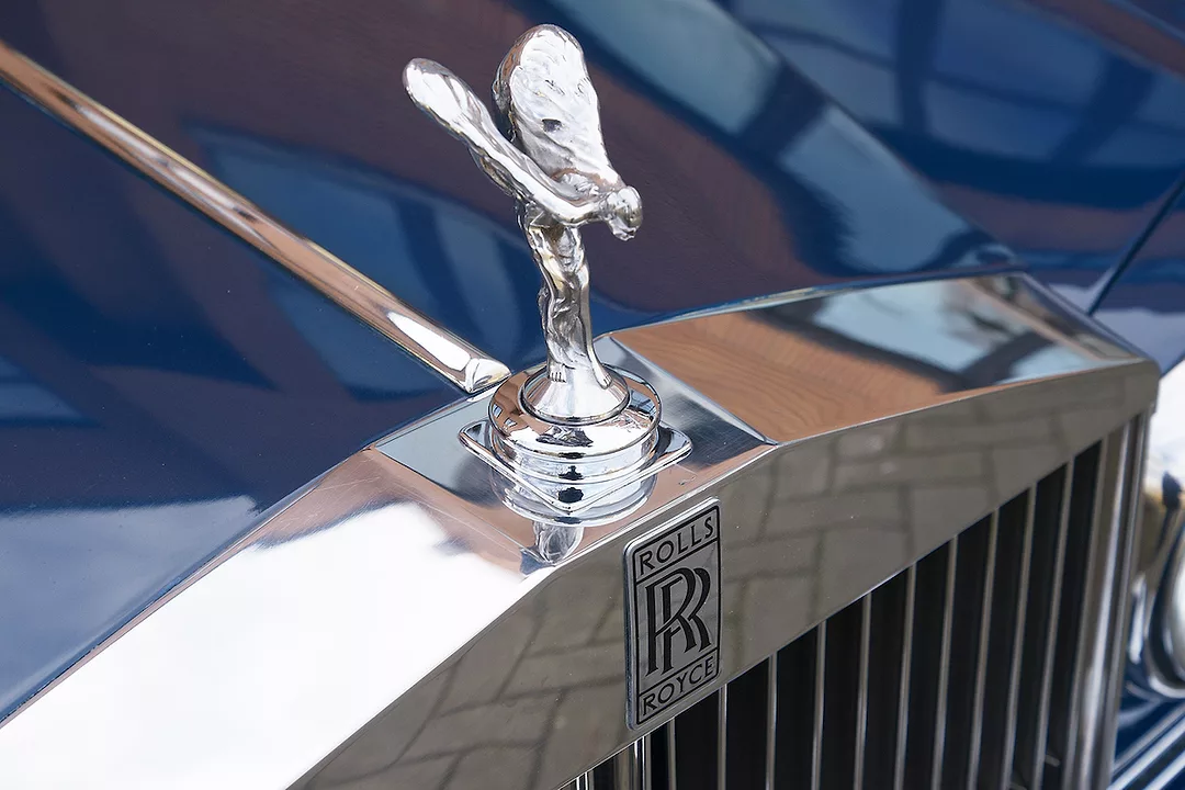 THE WINGED MASCOT OF ROLLS-ROYCE CAN BE SEEN ON THE HOOD OF A CAR WITH THE ROLLS-ROYCE LOGO RIGHT BELOW THE FIGURE.