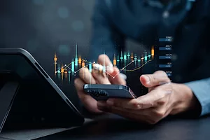 A PERSON HOLDING A CELL PHONE IN FRONT OF A TABLET. A DIGITAL RENDERING OF STOCK MARKET GRAPHS HOVERS ABOVE THE CELL PHONE.