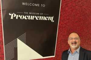 A MAN STANDS SMILING IN FRONT A SIGN THAT READS WELCOME TO THE MUSEUM OF PROCUREMENT