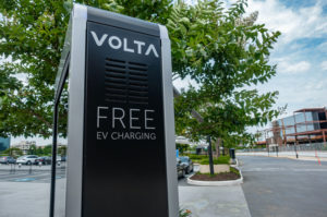 A BLACK AND SILVER VOLTA TRUCKS ELECTRIC VEHICLE CHARGER SITS IN A PARKING LOT. THE CHARGER READS "FREE EV CHARGING."