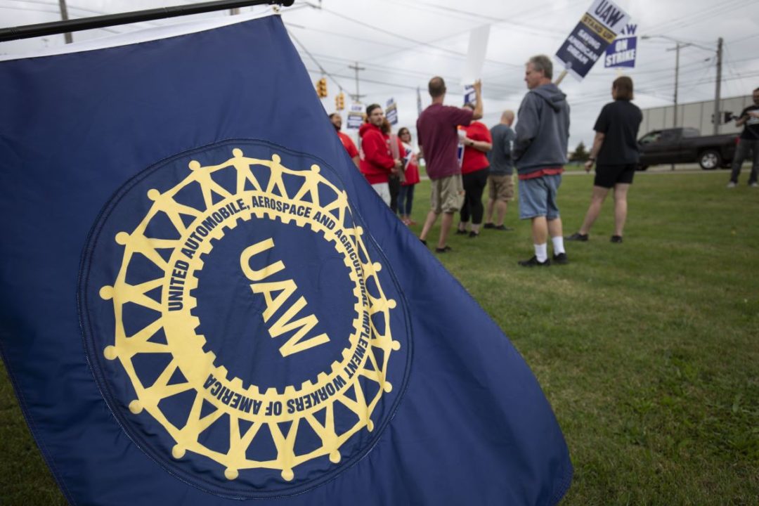 A BLUE AND GOLD UAW FLAG DOMINATES THE FOREGROUND, BLURRED IMAGES OF STRIKERS IN THE BACKGROUND