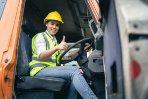A WOMAN SITTING IN THE DRIVER'S SEAT OF A TRUCK WEARING A YELLOW HELMET AND VEST GIVES A THUMBS UP TO THE CAMERA.