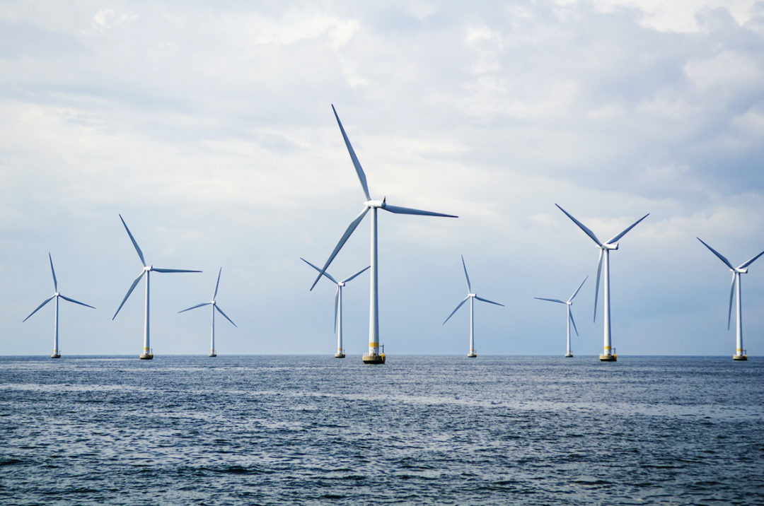 NINE WIND TURBINES CAN BE SEEN AT SEA UNDER A CLOUDY BLUE SKY.