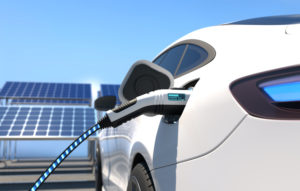 A CHARGER IS PLUGGED INTO THE LEFT SIDE OF A WHITE ELECTRIC VEHICLE. SOLAR PANELS CAN BE SEEN IN THE BACKGROUND UNDER A BLUE SKY.