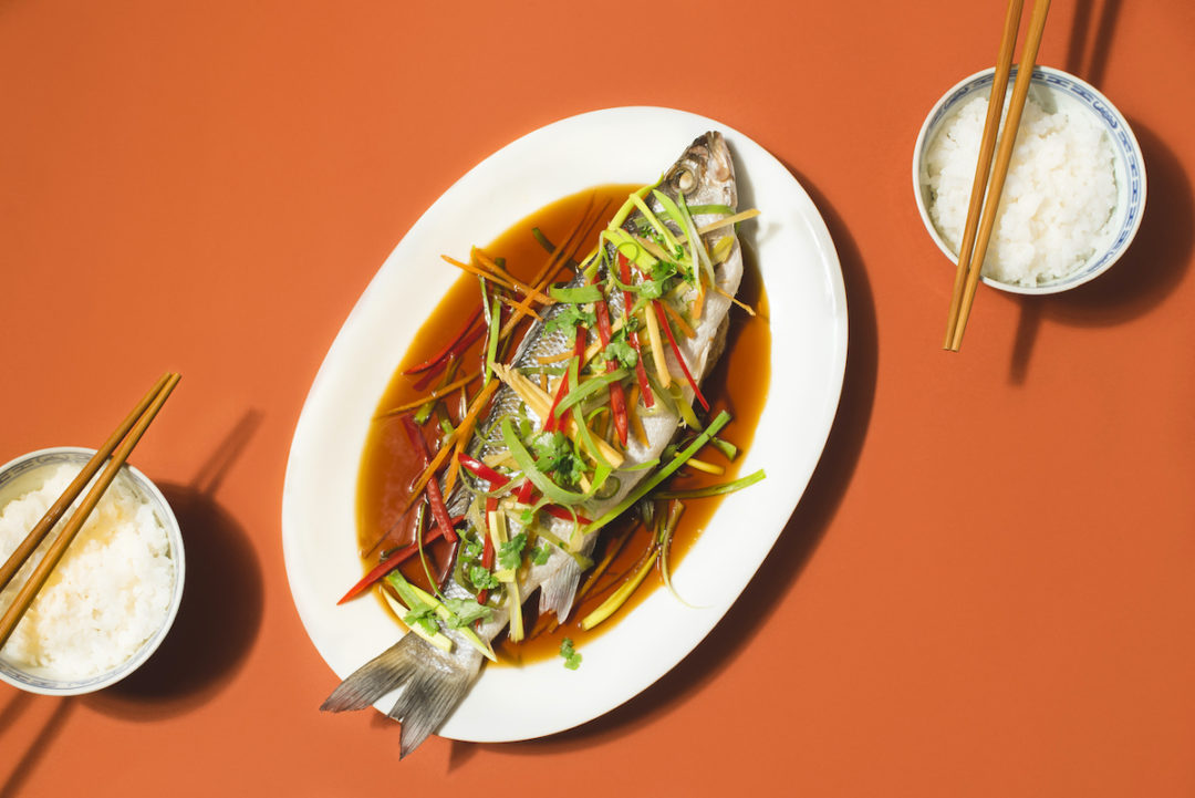 A COOKED FISH SITS ON AN OVAL PLATE ON TOP OF SOY SAUCE UNDERNEATH A BED OF VEGETABLES. TWO BOWLS OF RICE WITH CHOPSTICKS ON TOP OF THEM SIT ON OPPOSITE SIDES OF THE PLATE IN FRONT OF AN ORANGE BACKGROUND.