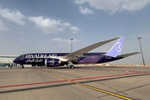 A DARK BLUE PLANE WITH THE WORDS RIYADH AIR ON THE SIDE SITS ON TARMAC AT AN AIRPORT