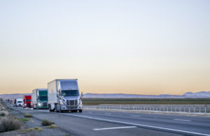 A FLEET OF LONG HAULER SEMI TRUCKS ARE DRIVING ON AN EMPTY HIGHWAY IN A LINE.