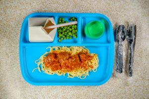 A PLASTIC TRAY WITH DIVIDED SECTIONS HOLDS MEATBALLS AND SPAGETTI, PEAS, A GLASS OF WATER AND AN OPEN SMALL CARTON WITH A STRAW IN IT.