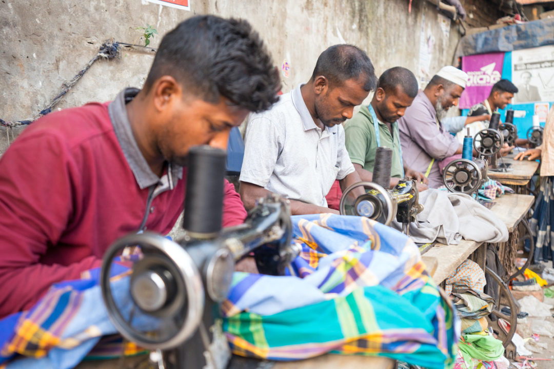 A GROUP OF TAILORS CAN BE SEEN WORKING AT SEWING MACHINES.