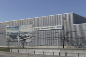 EXTERIOR OF A FORD ASSEMBLY PLANT. NEXT TO A LARGE IMAGE OF FORD VEHICLES IS A SIGN THAT READS "CHICAGO ASSEMBLY PLANT" FOLLOWED BY THE FORD LOGO.